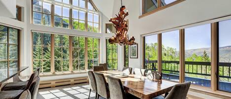 A spacious dining area with a large wooden table and chairs, large windows offering views of greenery and mountains, and a unique chandelier.