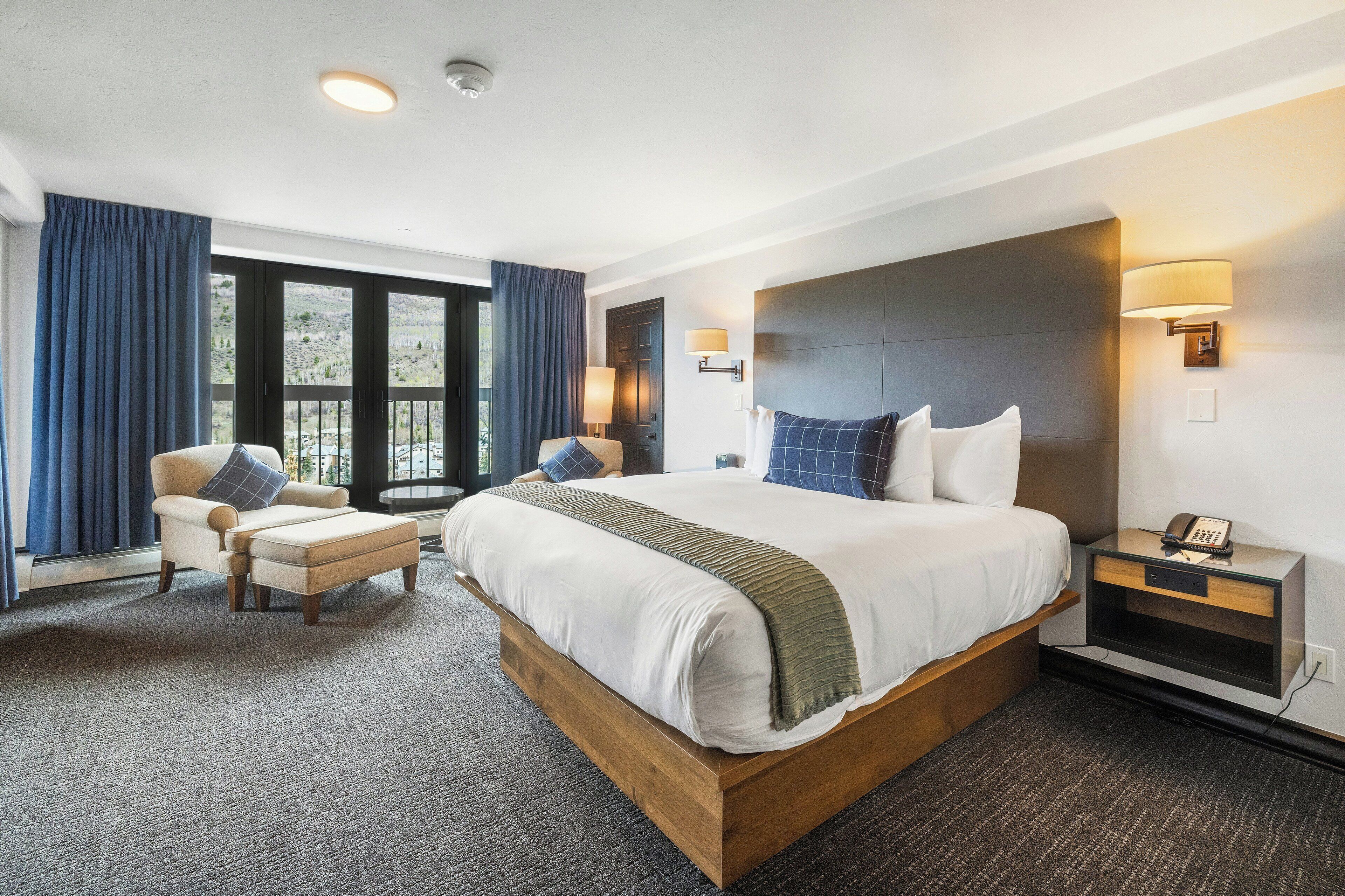 A hotel room with a large bed, a chair, a bedside table with a lamp, and a view of the outdoors.