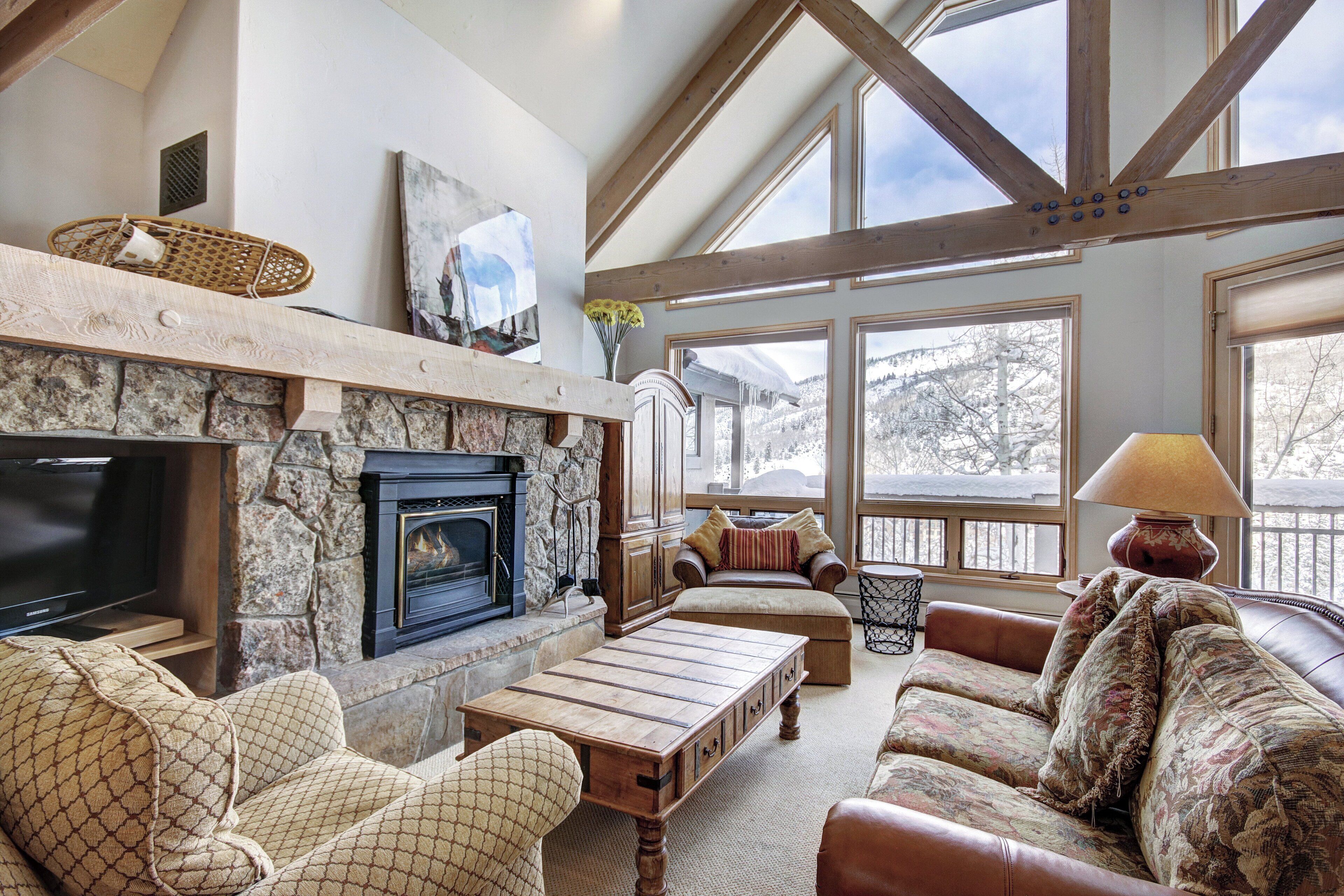 A cozy living room with a stone fireplace, wooden beams, a television, and a large window overlooking a snowy landscape.