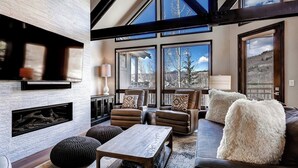 Una sala de estar moderna con chimenea, sofás de cuero, una mesa de centro y ventanas amplias que ofrecen vista a árboles y montañas.