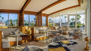A spacious outdoor dining area with multiple tables and chairs, large windows offering views of palm trees and buildings, and wooden architectural details. - Bucuti & Tara Beach Resort - Adults Only (Oranjestad)