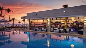 Zona de piscina con restaurante, sillas de descanso, palmeras y vista al atardecer.