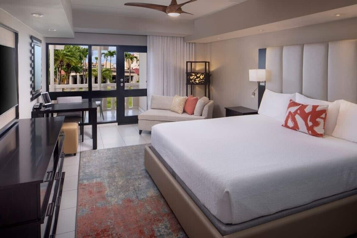 A modern hotel room with a large bed, a desk, a sofa, and a view of palm trees.