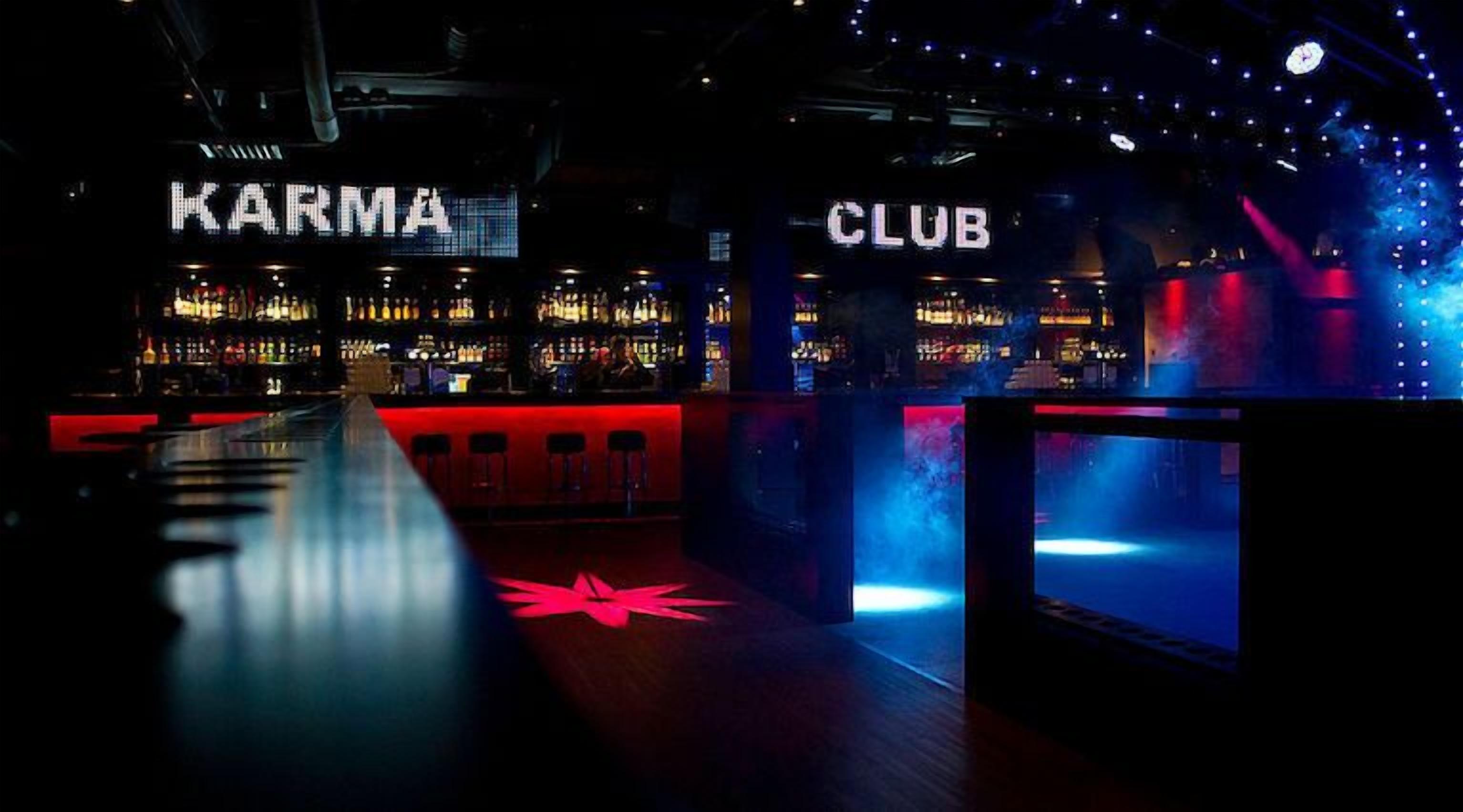 Zona de bar con letreros que dicen 'KARMA' y 'CLUB', mostradores de bar y suelo reflectante.