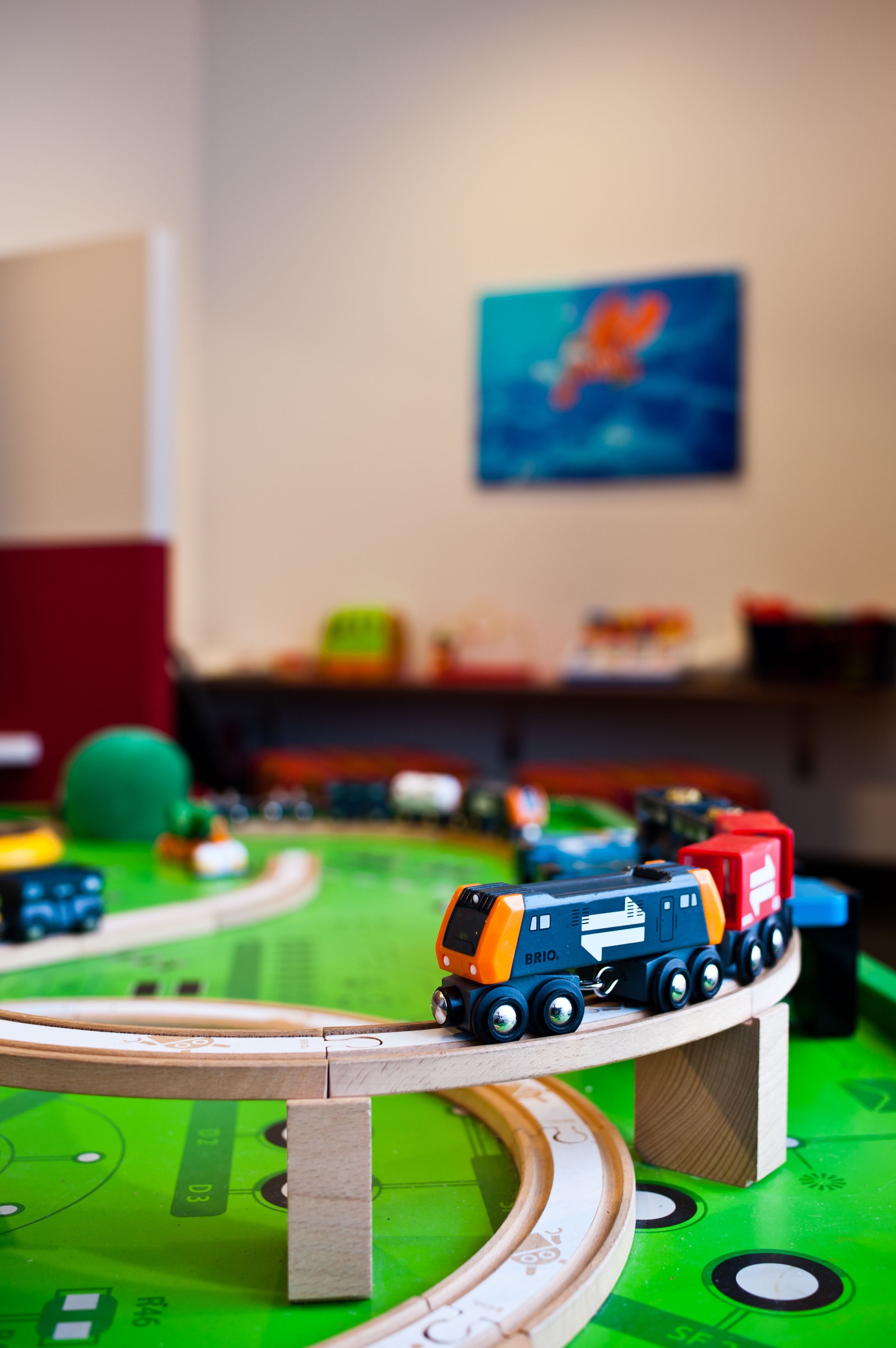 A toy train set on a green track with a wooden bridge, surrounded by various toys and a painting on the wall.