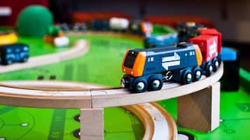 A toy train set on a green track with a wooden bridge, surrounded by various toys and a painting on the wall.
