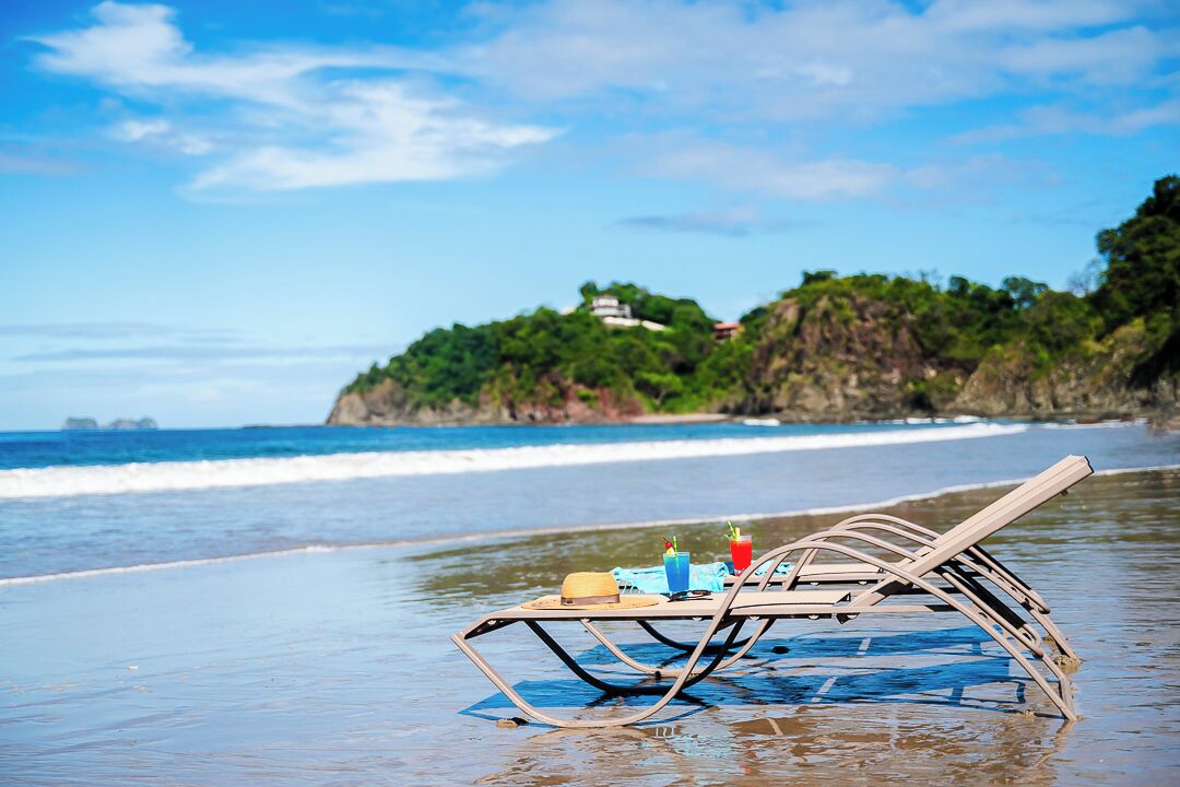 Escena de playa con una tumbona y una mesa dispuestas sobre la arena, un sombrero y dos bebidas sobre la mesa.