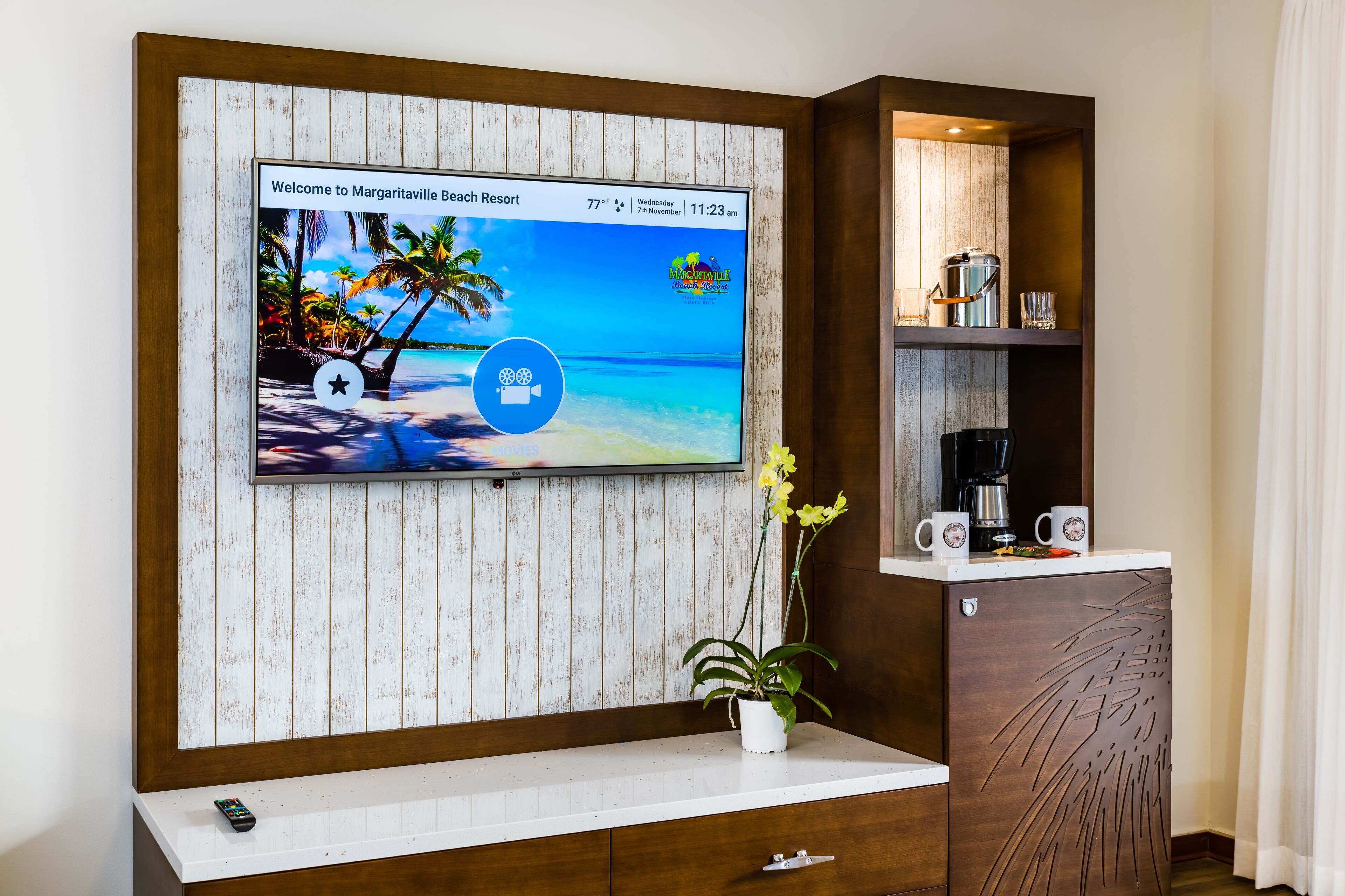 Un espace d’accueil d’hôtel avec un téléviseur diffusant des images d’une plage tropicale, une machine à café et une plante posée sur le comptoir.