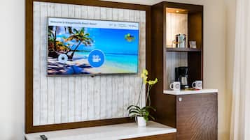 Un espace d’accueil d’hôtel avec un téléviseur diffusant des images d’une plage tropicale, une machine à café et une plante posée sur le comptoir.