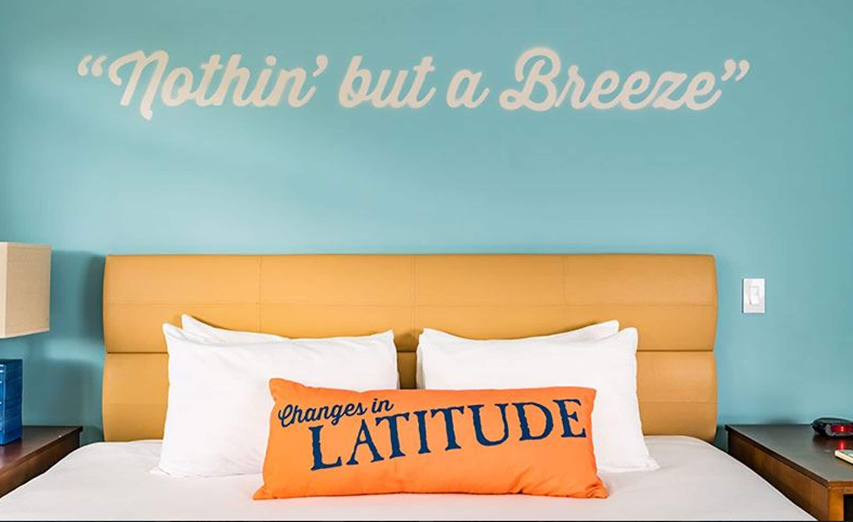 A bed with white pillows and an orange pillow with "Changes in LATITUDE" text, against a teal wall with "Nothin' but a Breeze" text.