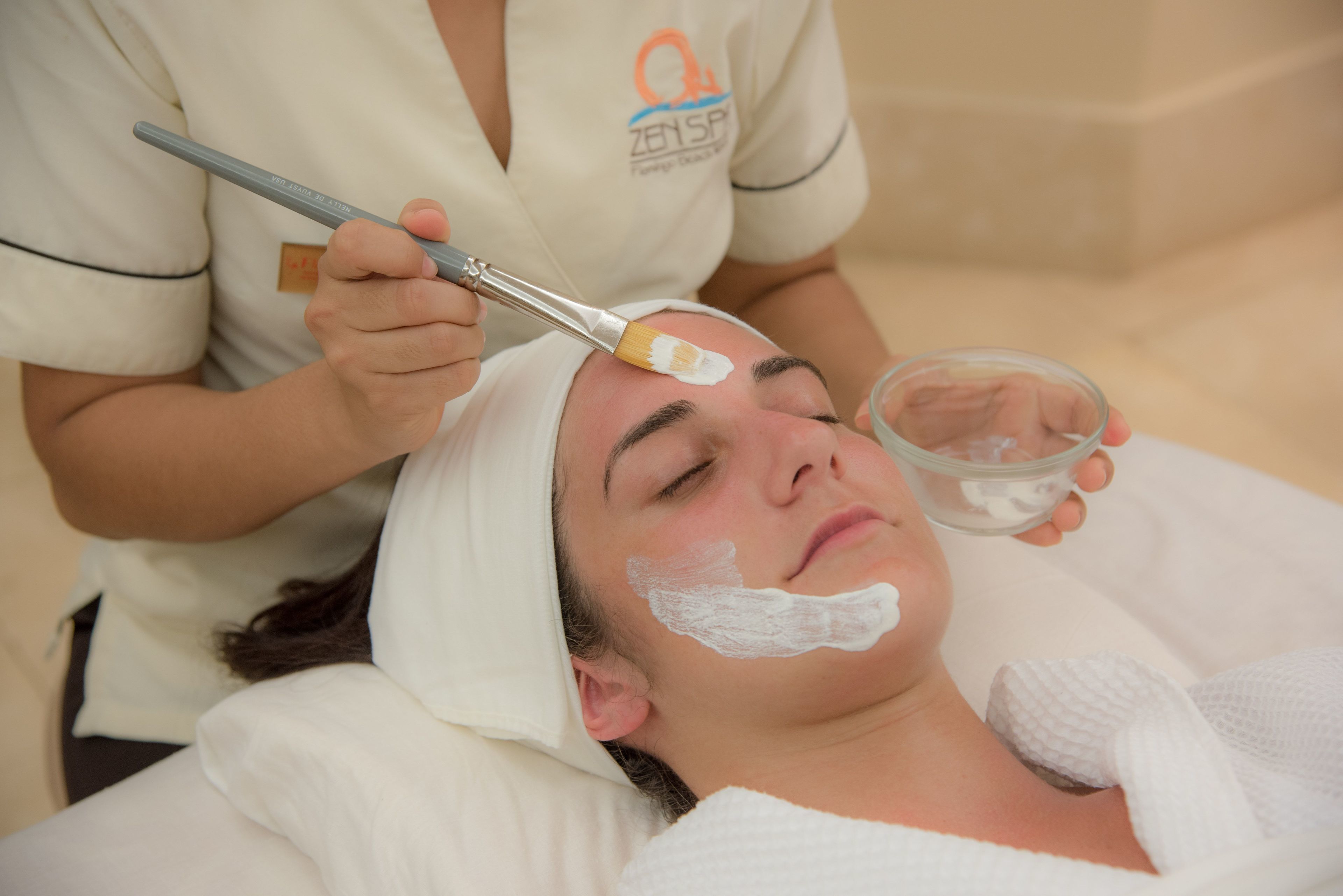 Una persona recibiendo un tratamiento facial con un cepillo y un cuenco de crema.