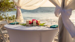 Taula rodona preparada per a dinar a la platja, amb estovell blanc, espelmes i flors.