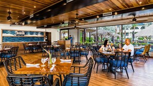 A spacious restaurant with wooden flooring and high ceilings, featuring a bar area and outdoor seating with a view of the pool.