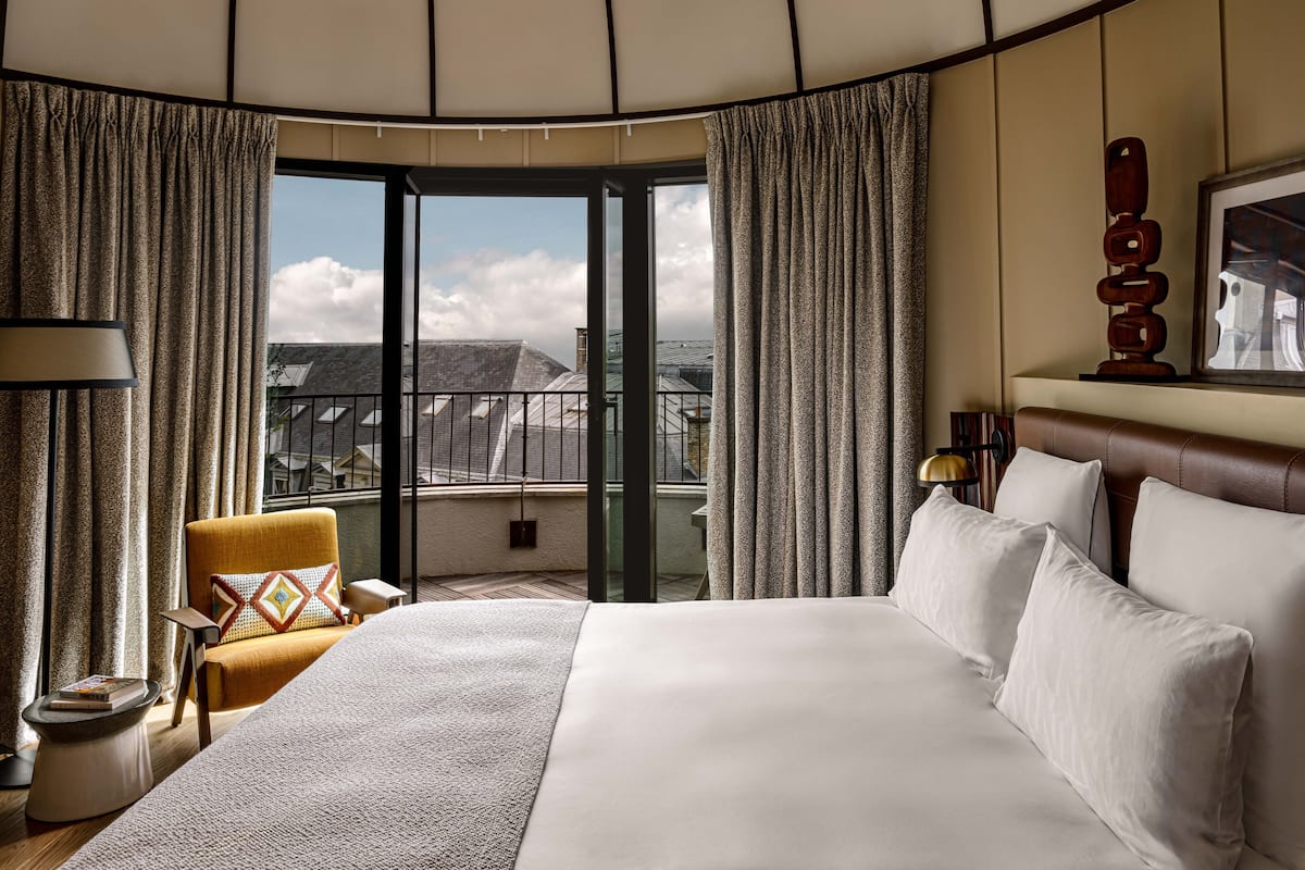A hotel room with a large bed, a yellow armchair, a view of buildings through a glass door, and a wooden sculpture on the wall.
