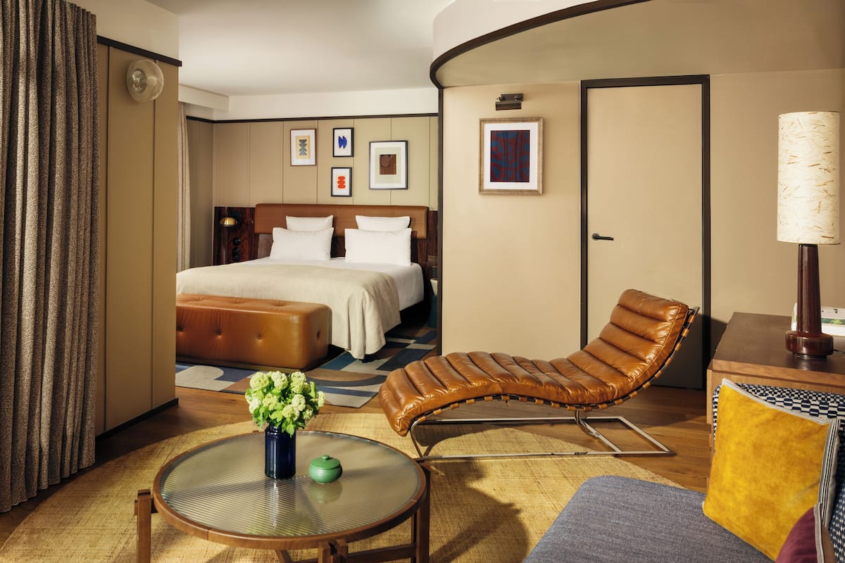 A hotel room with a bed, a lounge chair, a small table with a vase, and a lamp.