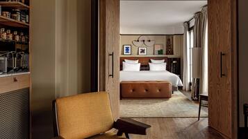 A hotel room with a bed, a chair, a wooden cabinet with kitchen appliances, and a view into another room.