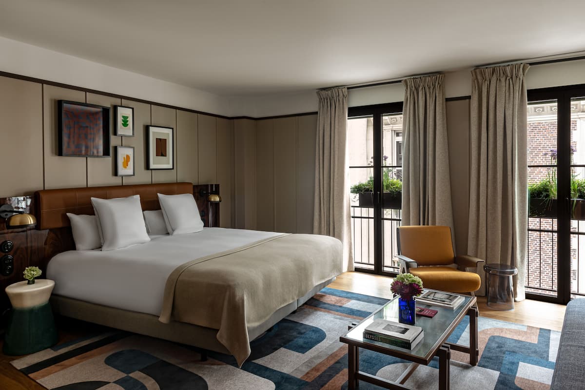 A hotel room with a large bed, a side table, a chair, a coffee table, and a view of the outside through a window with curtains.