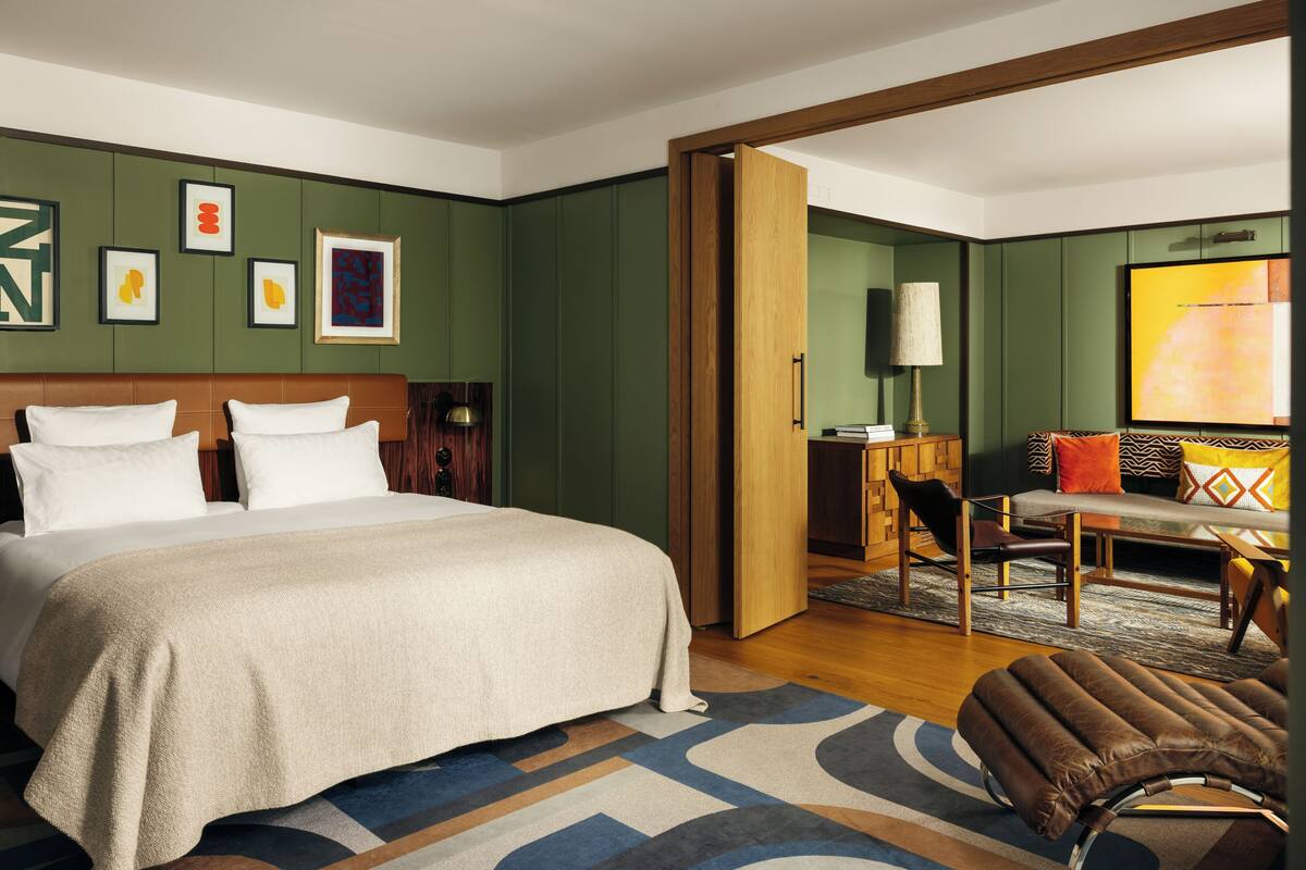 A hotel room with a large bed, a desk, a chair, and a sofa. There are framed pictures on the wall and a TV on the wall.