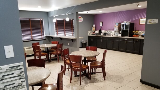 A dining area with round tables and chairs, a kitchen area with cabinets and appliances, and a sign indicating hours of operation. - Super 8 by Wyndham Fort Collins (Fort Collins)