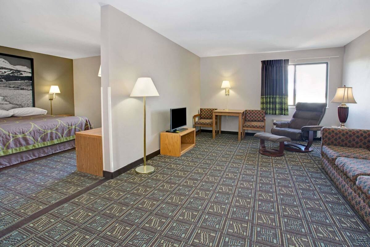 A hotel room with a bed, a desk with a TV, a sofa, two chairs, a patterned carpet, and a window with curtains.
