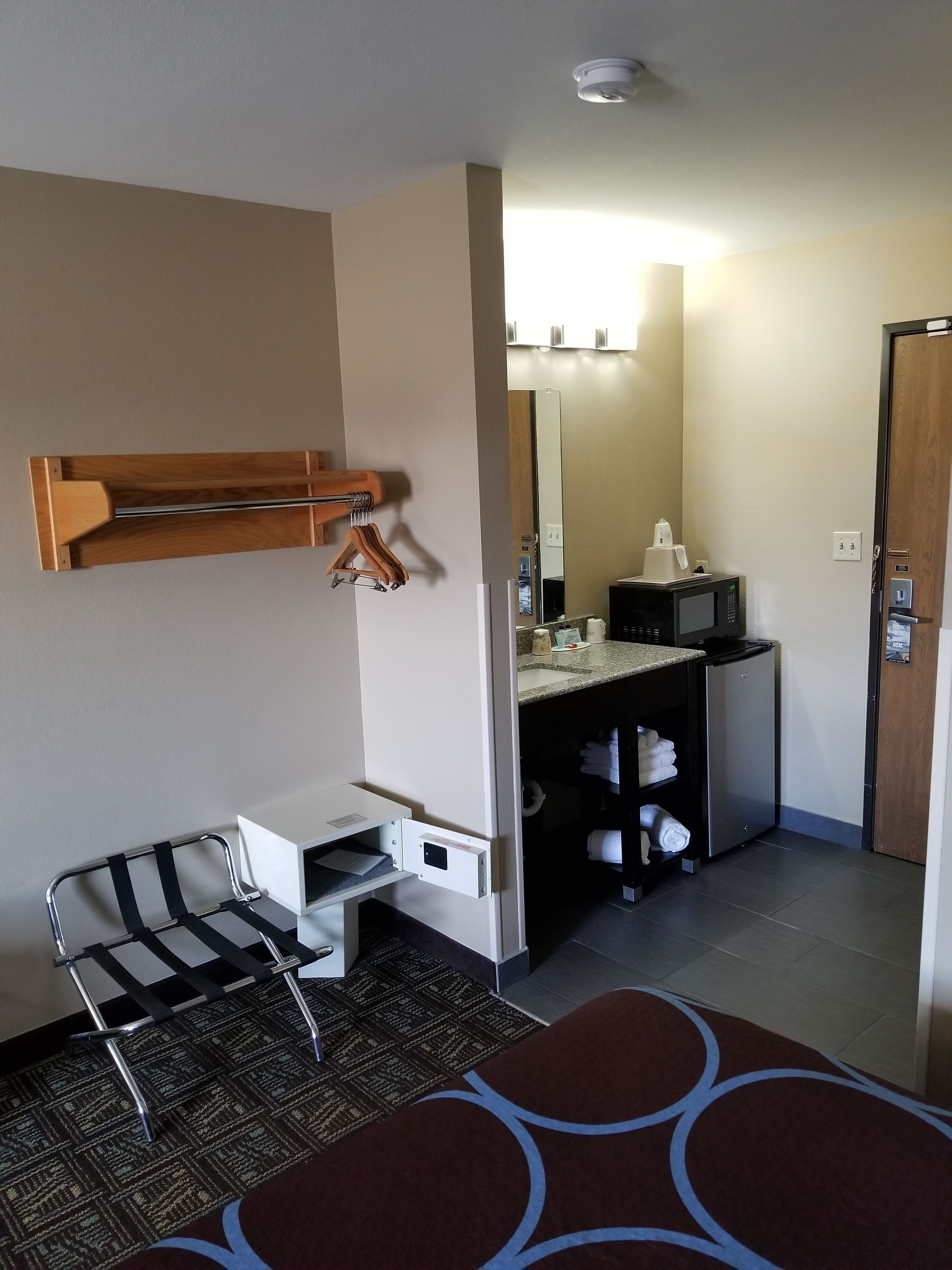 A hotel room with a bed, a desk with a chair, a microwave, a small refrigerator, a mirror, and a door.