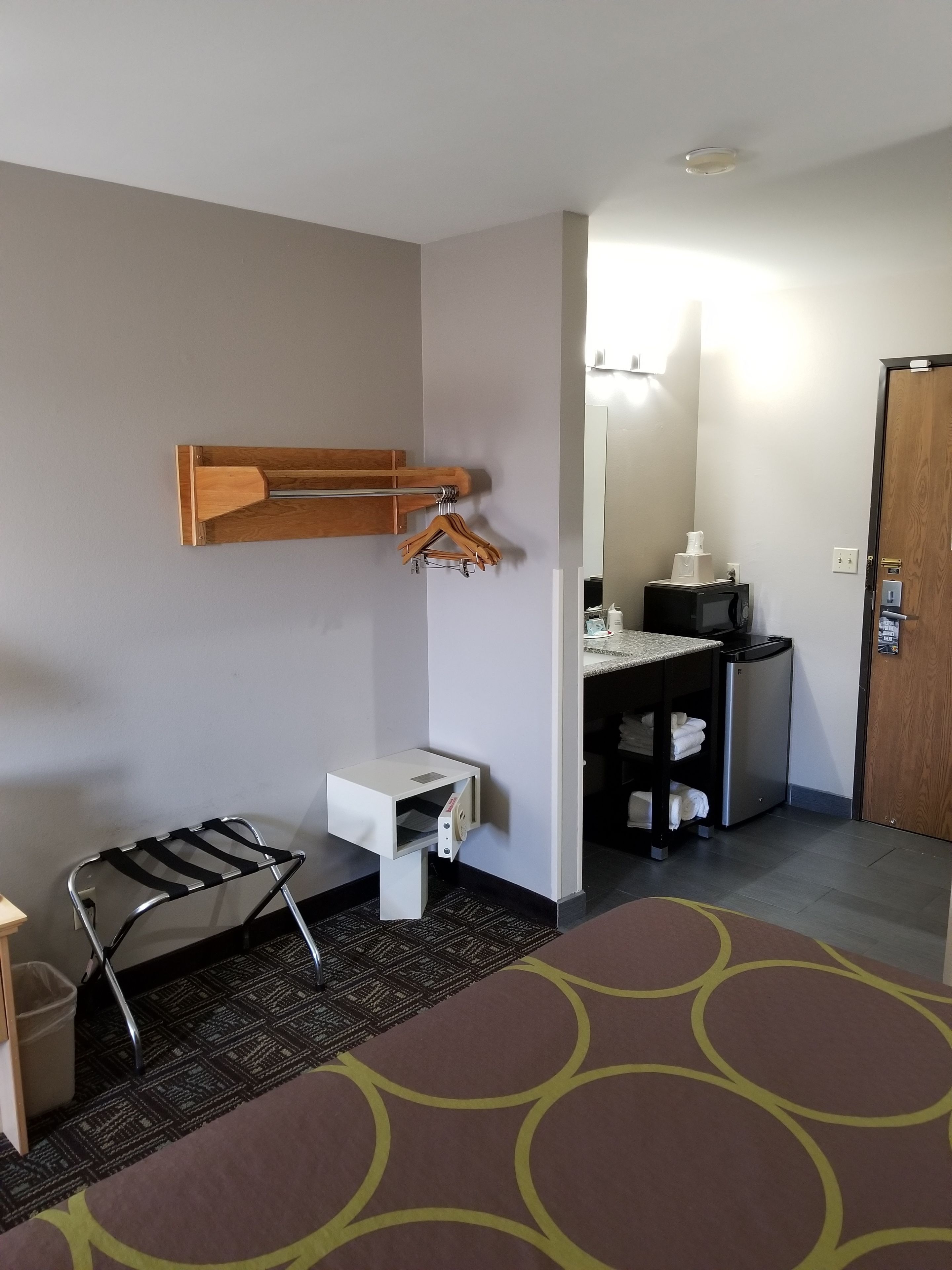 Quarto de hotel com uma cama, uma pequena mesa, uma cadeira, um cabideiro de madeira, micro-ondas, pia e porta.