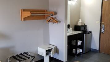 Quarto de hotel com uma cama, uma pequena mesa, uma cadeira, um cabideiro de madeira, micro-ondas, pia e porta.