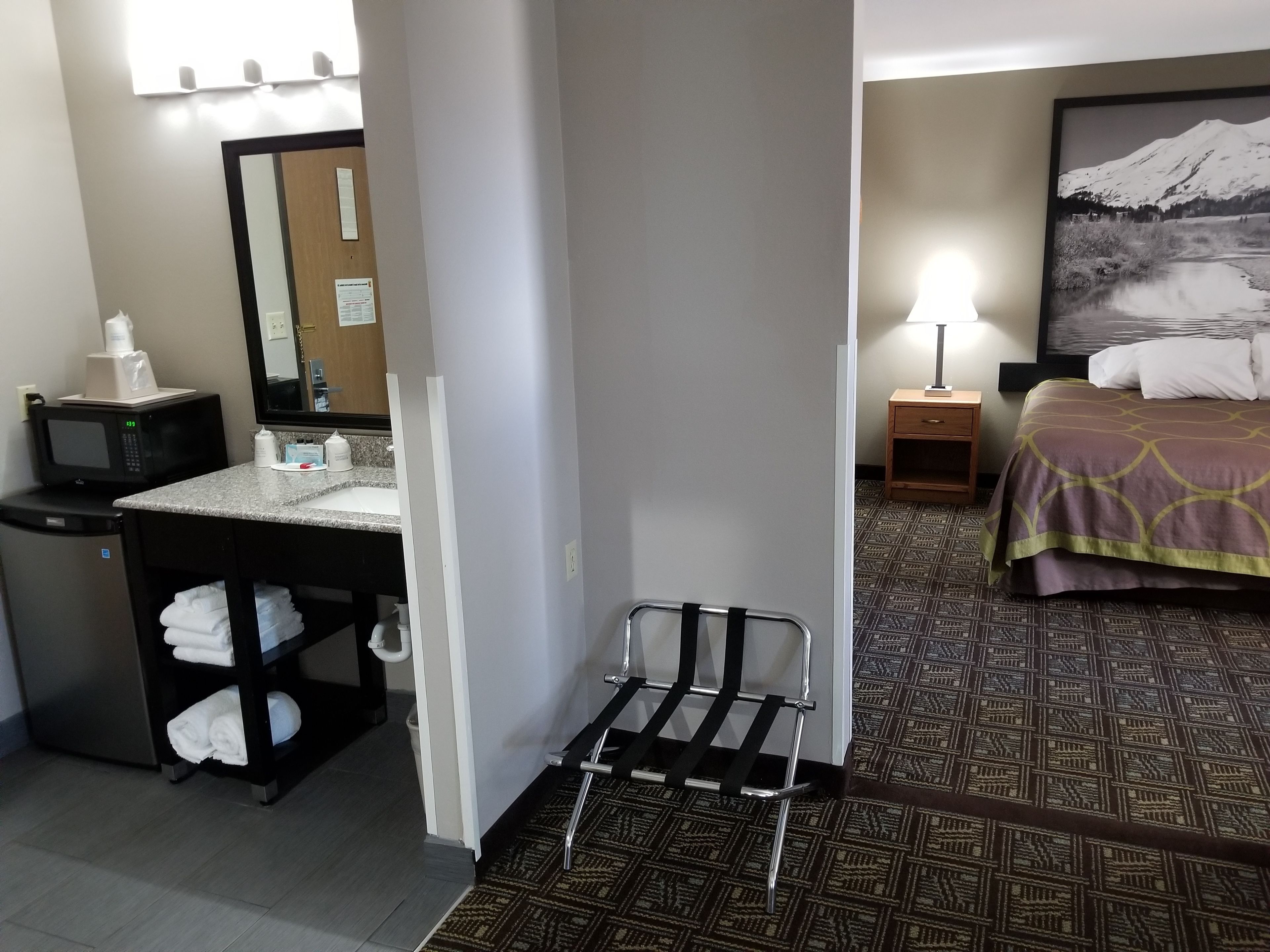 A hotel room with a bathroom, a bed with a patterned bedspread, a nightstand, and a painting of a mountain.