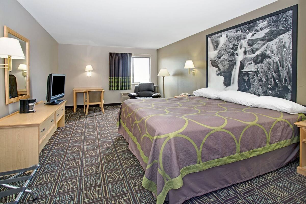 A hotel room with a bed, a desk, a chair, a television, and a large framed picture on the wall.