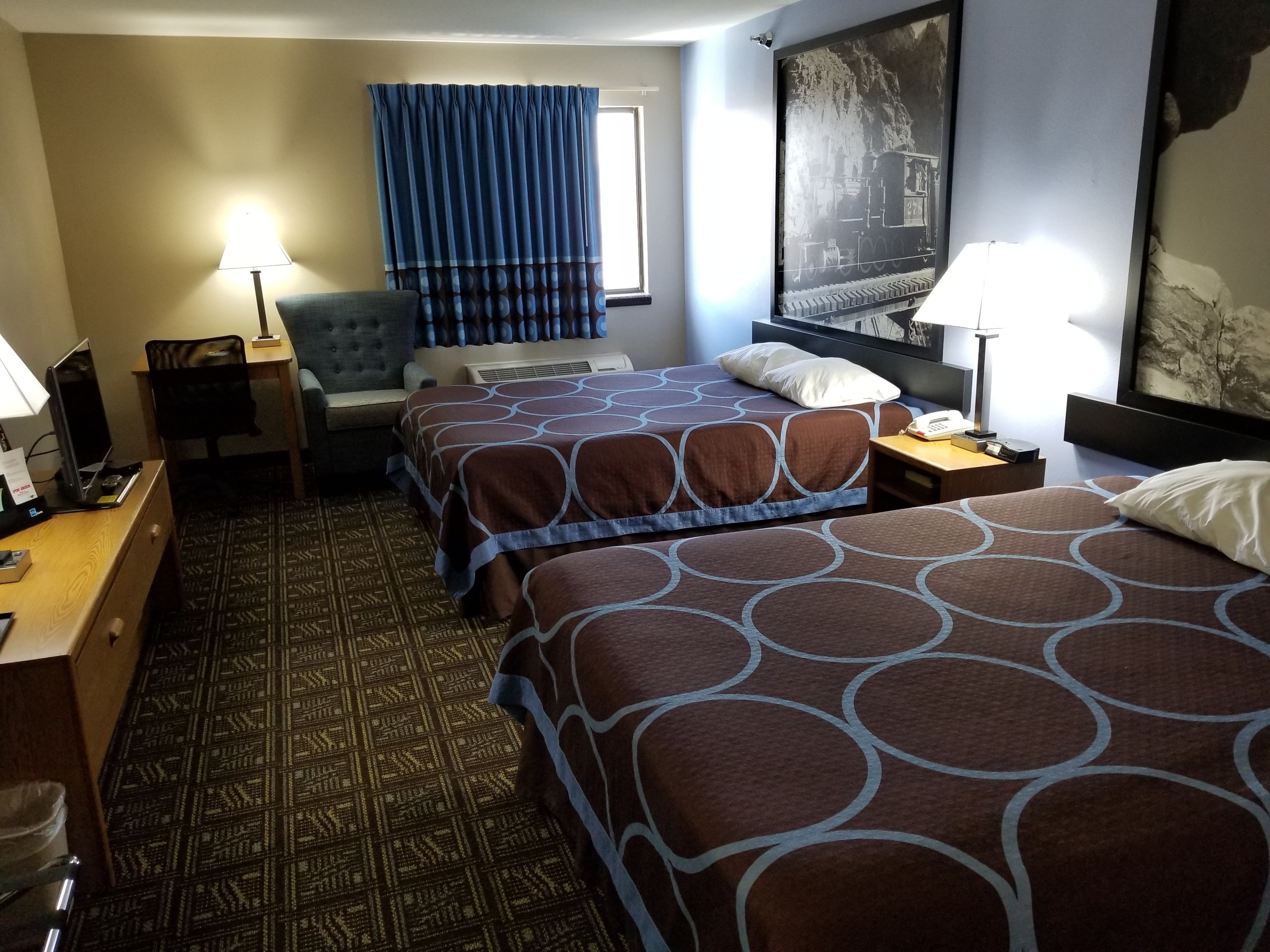 A hotel room with two beds, a desk, a chair, a lamp, and a large painting on the wall.