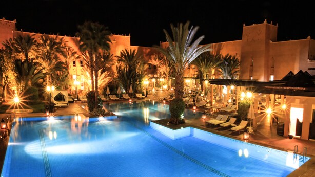 A nighttime swimming pool area with lounge chairs and palm trees, surrounded by buildings with warm lighting. - Le Berbere Palace (Ouarzazate)