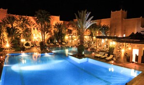 A nighttime swimming pool area with lounge chairs and palm trees, surrounded by buildings with warm lighting. - Le Berbere Palace (Ouarzazate)
