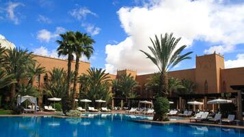 A pool area with lounge chairs and umbrellas, surrounded by palm trees and a building with a beige facade.