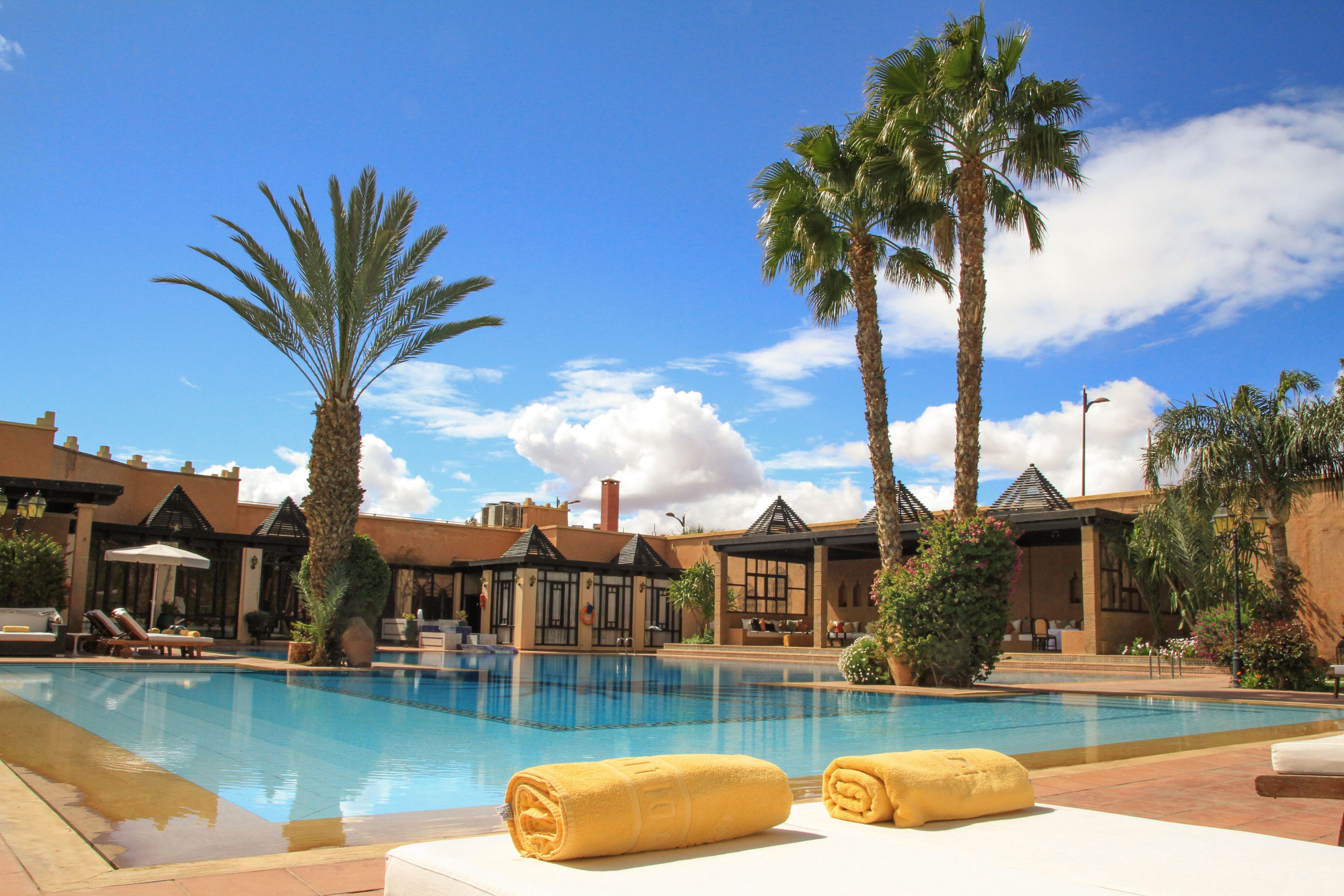 A swimming pool with lounge chairs and towels, surrounded by palm trees and a building with a tiled roof.