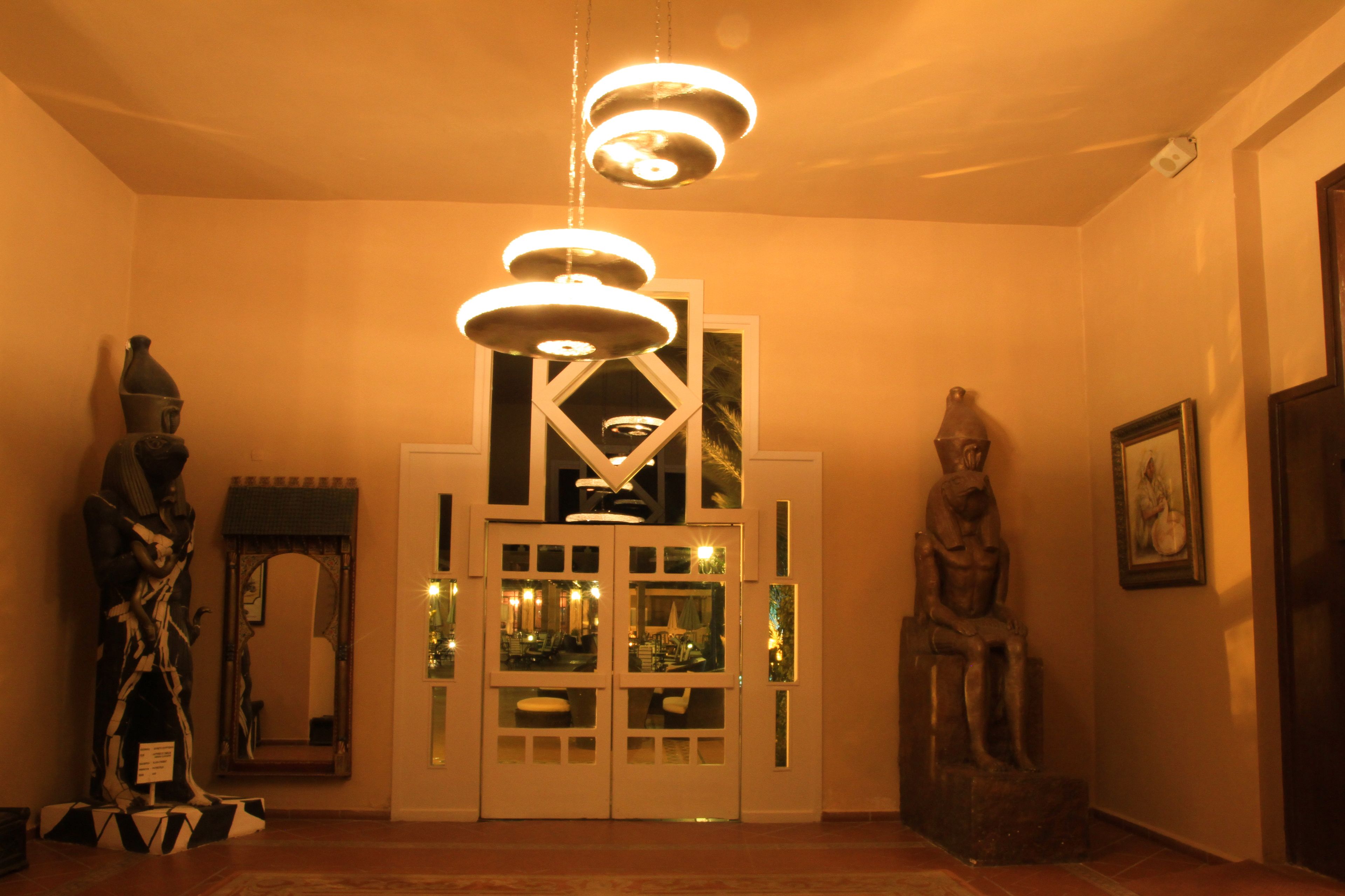 Un couloir chaleureux, éclairé par une lumière douce, avec un luminaire décoratif au plafond, un mur recouvert de miroirs et des statues de part et d’autre.
