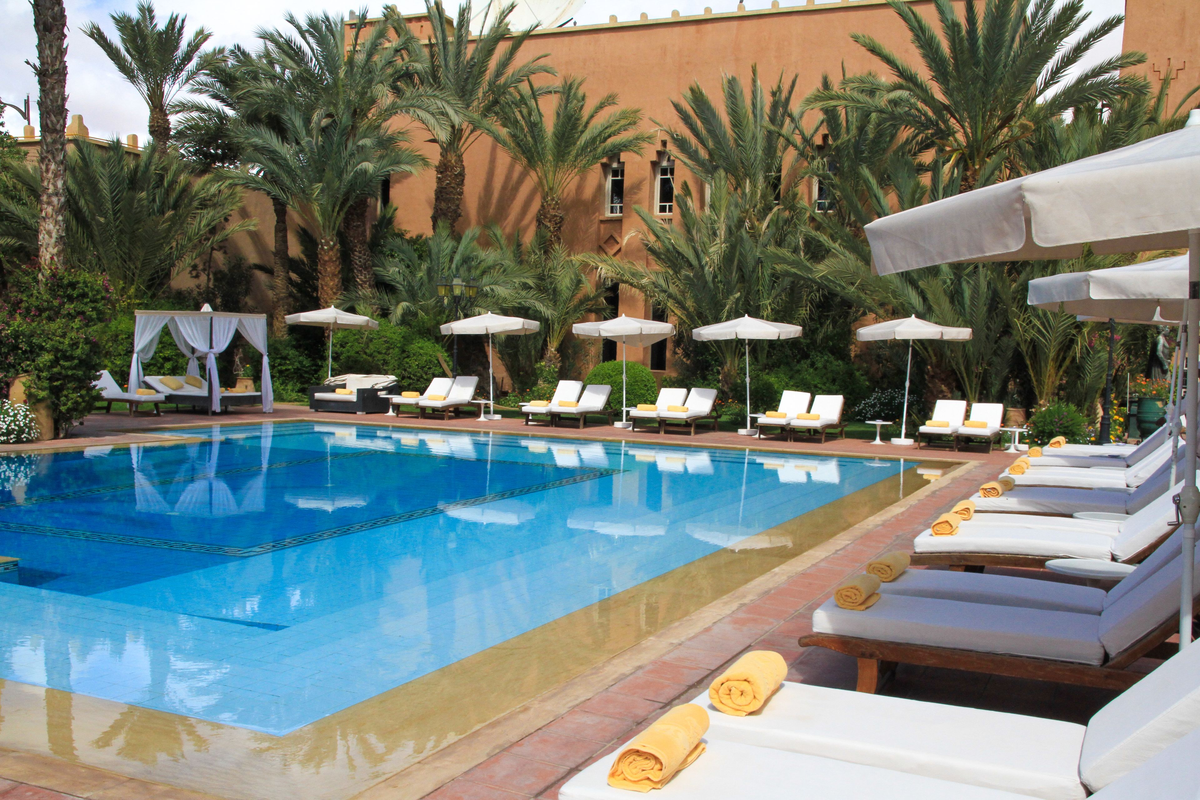 Una piscina con sillas de descanso y sombrillas, rodeada de palmeras y un edificio al fondo.