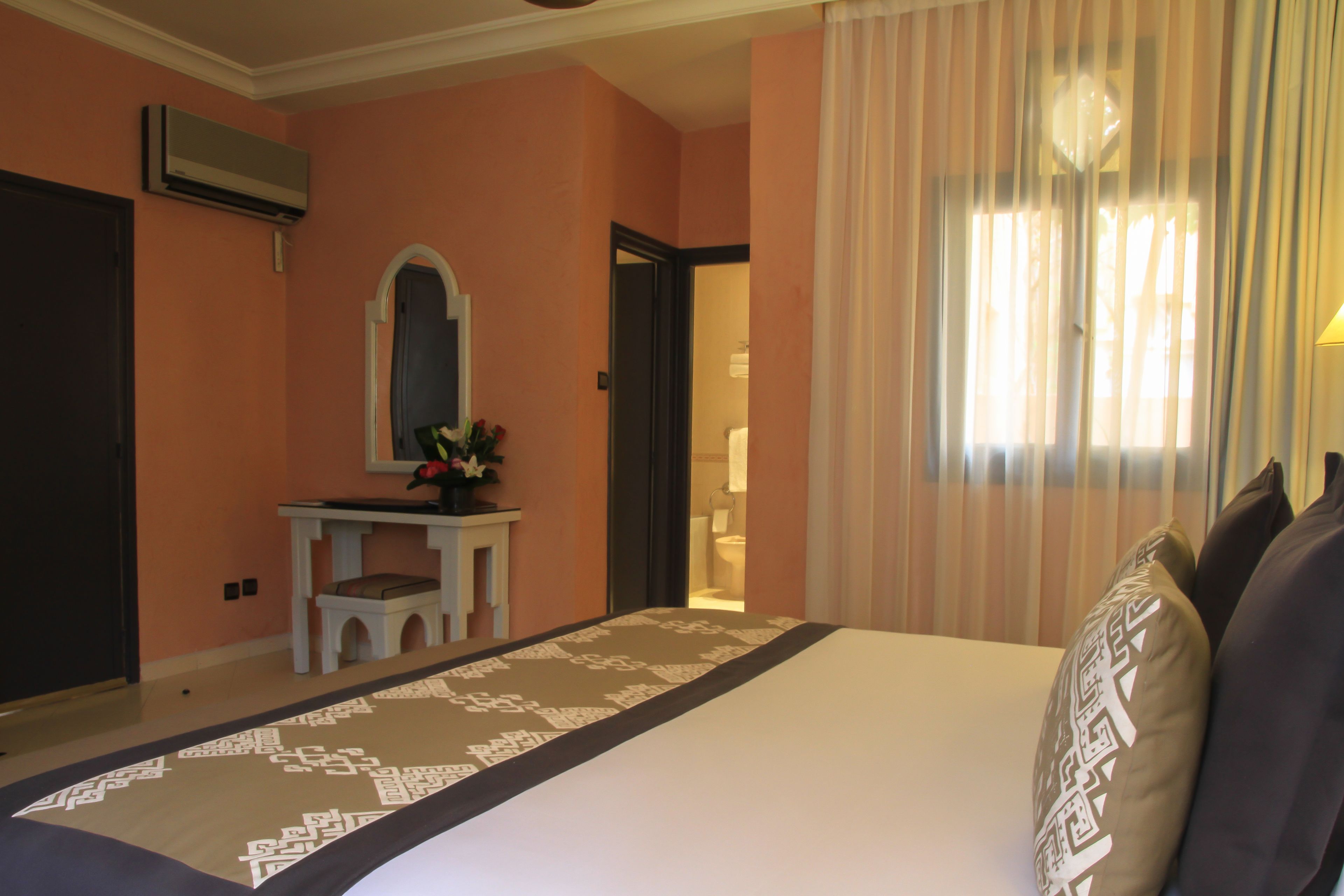 A hotel room with a large bed, a vanity with a mirror, and a window with sheer curtains.