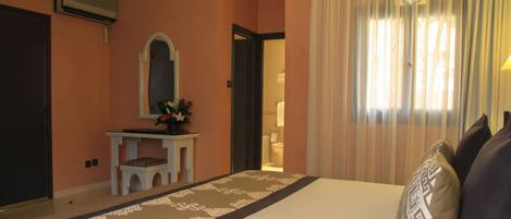 Una habitación de hotel con una cama grande, un tocador con espejo y una ventana con cortinas translúcidas.