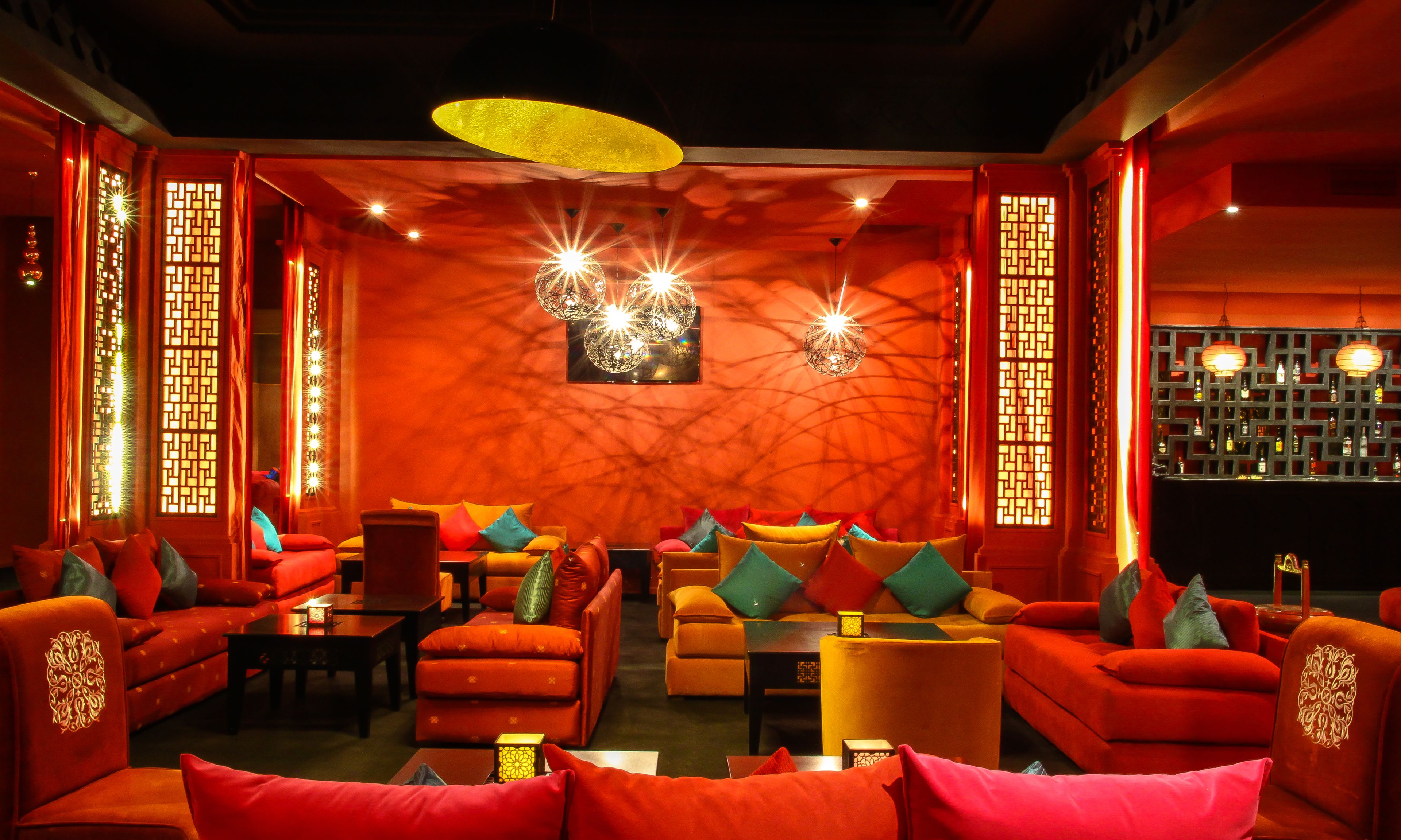 Un área de sala con luz cálida, sofás rojos, una barra con botellas e iluminación decorativa.