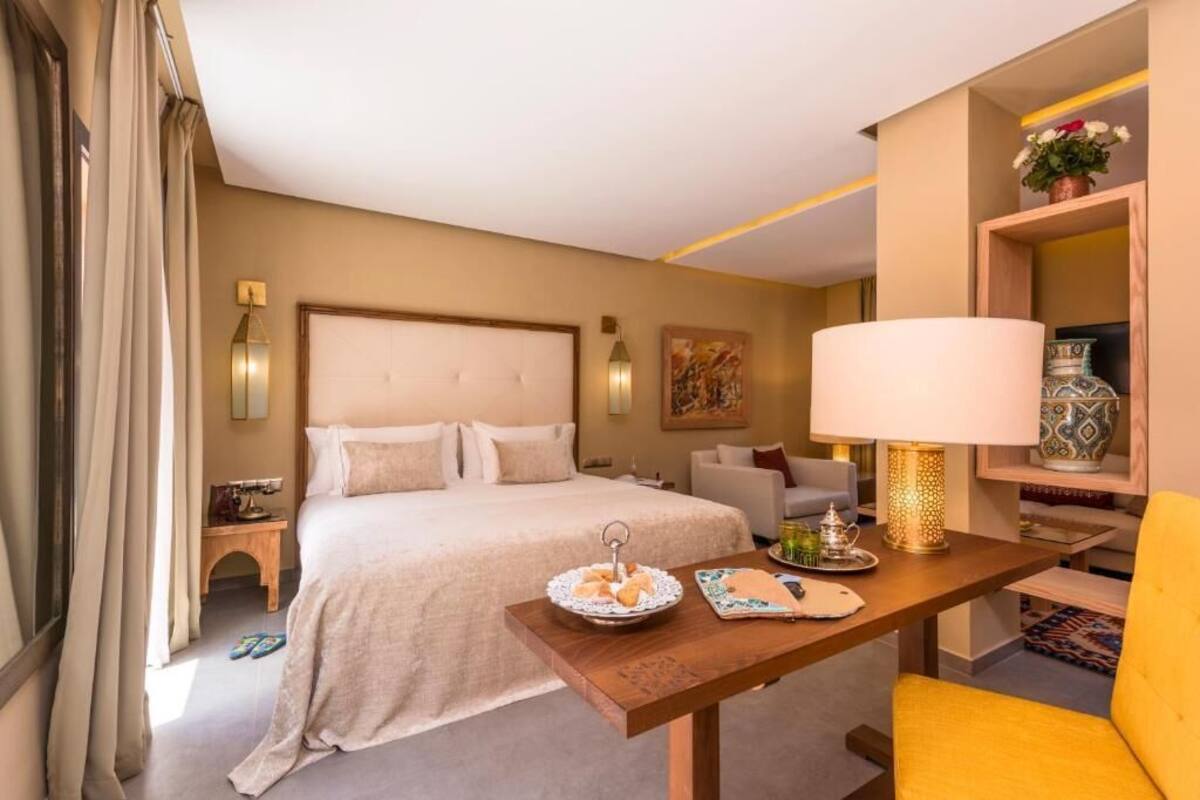 A hotel room with a bed, a desk with a lamp, a chair, a small table with a tray, and a shelf with decorative items.