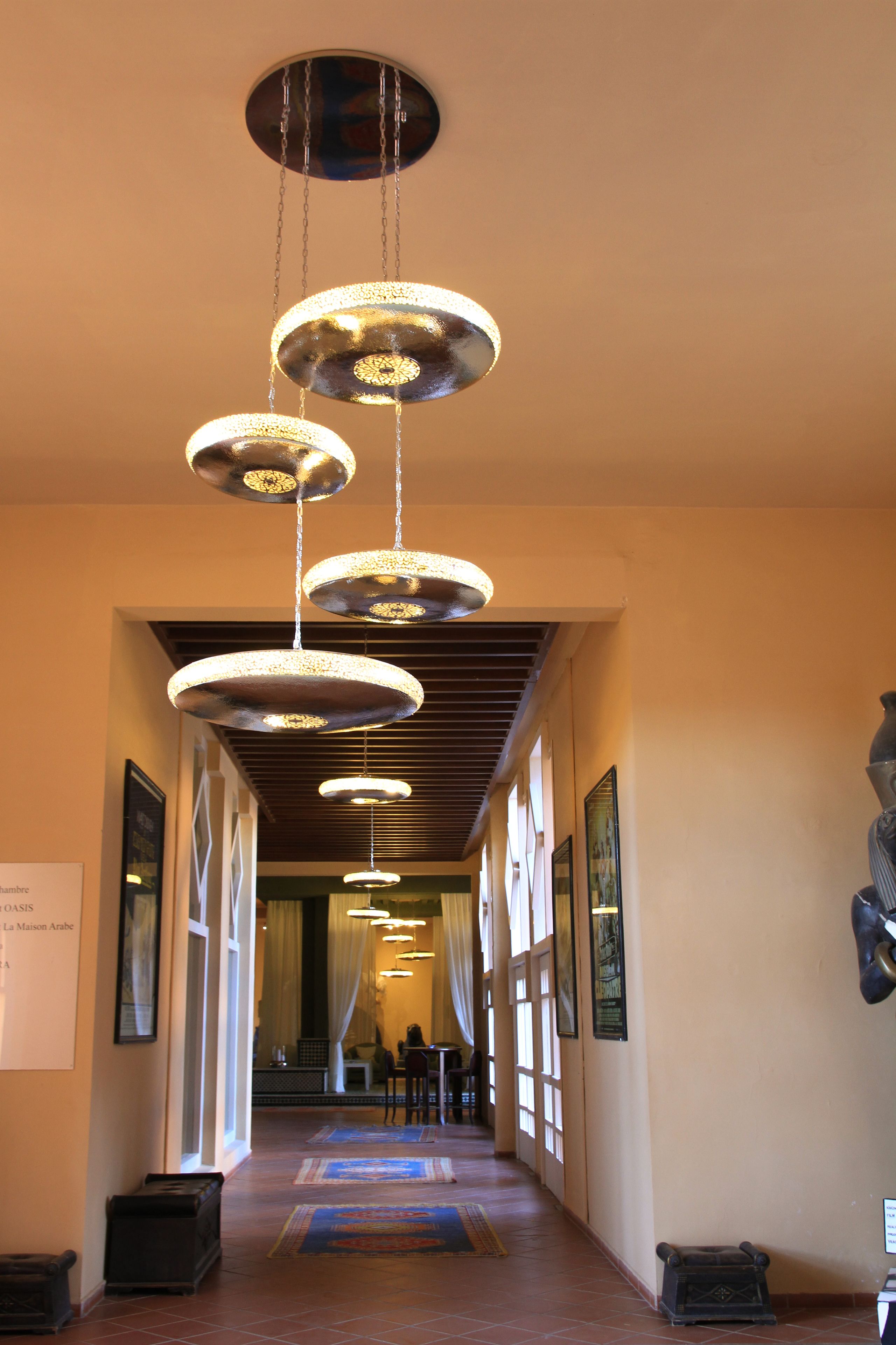 Un pasillo de hotel con lámparas circulares colgantes, suelo de baldosas y cuadros en las paredes.