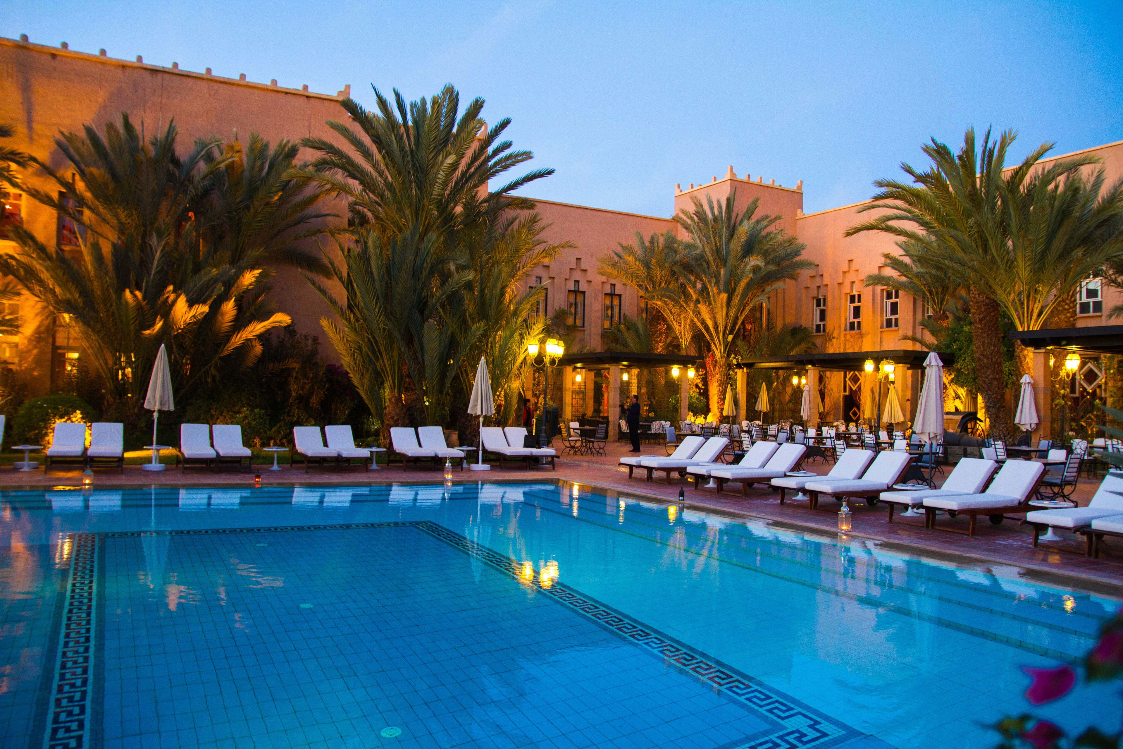 A swimming pool with lounge chairs and umbrellas, surrounded by palm trees and a building with warm lighting.