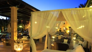 An outdoor lounge area with white canopies, wicker furniture, and a pool in the background. - Le Berbere Palace (Ouarzazate)