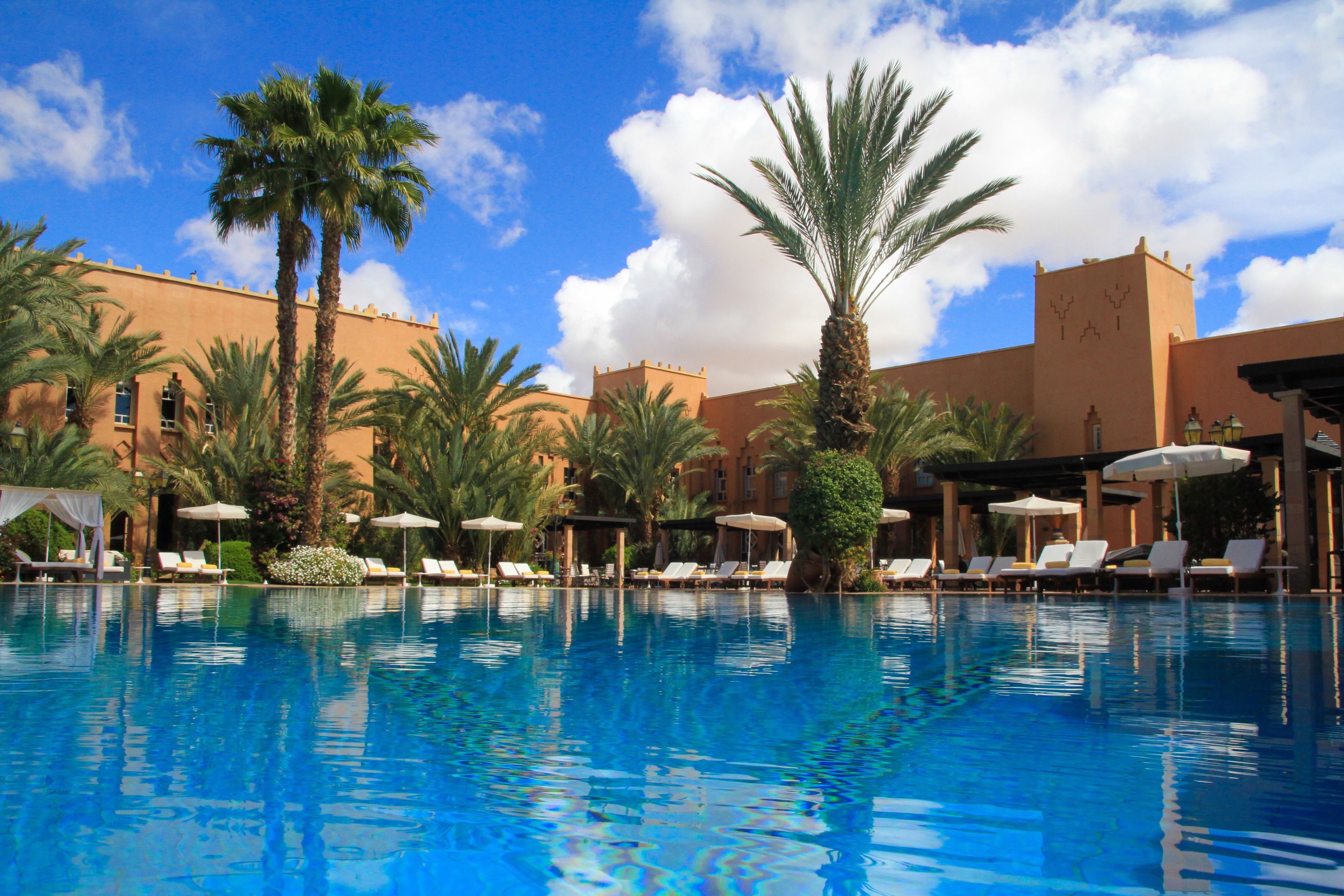 A swimming pool with lounge chairs and umbrellas, surrounded by palm trees and a building with a tower.
