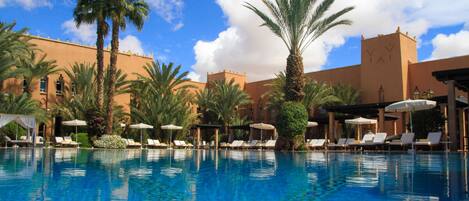 A swimming pool with lounge chairs and umbrellas, surrounded by palm trees and a building with a tower.