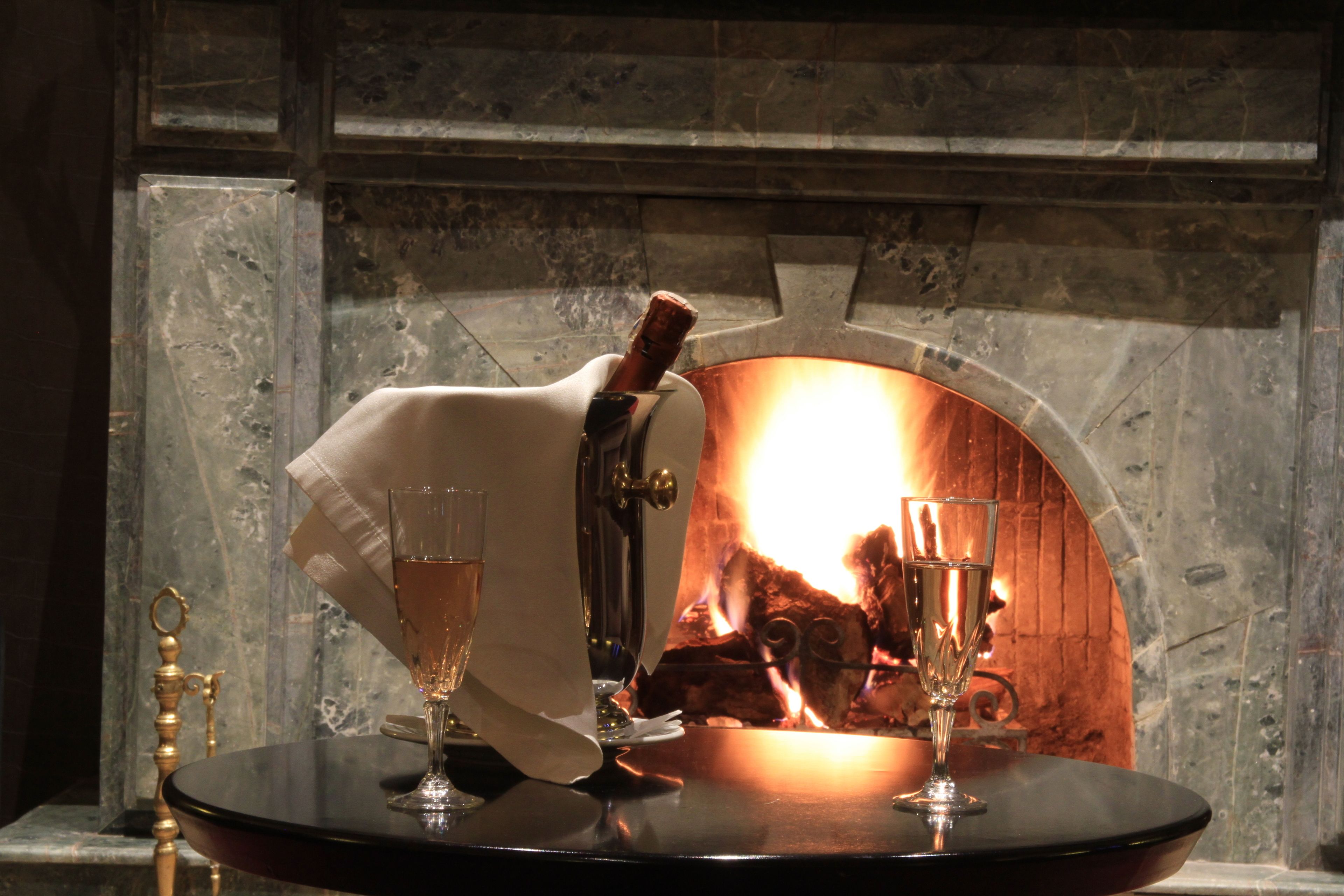 Ein Tisch mit zwei Sektgläsern und einer Flasche vor einem Steinkamin mit brennendem Feuer.