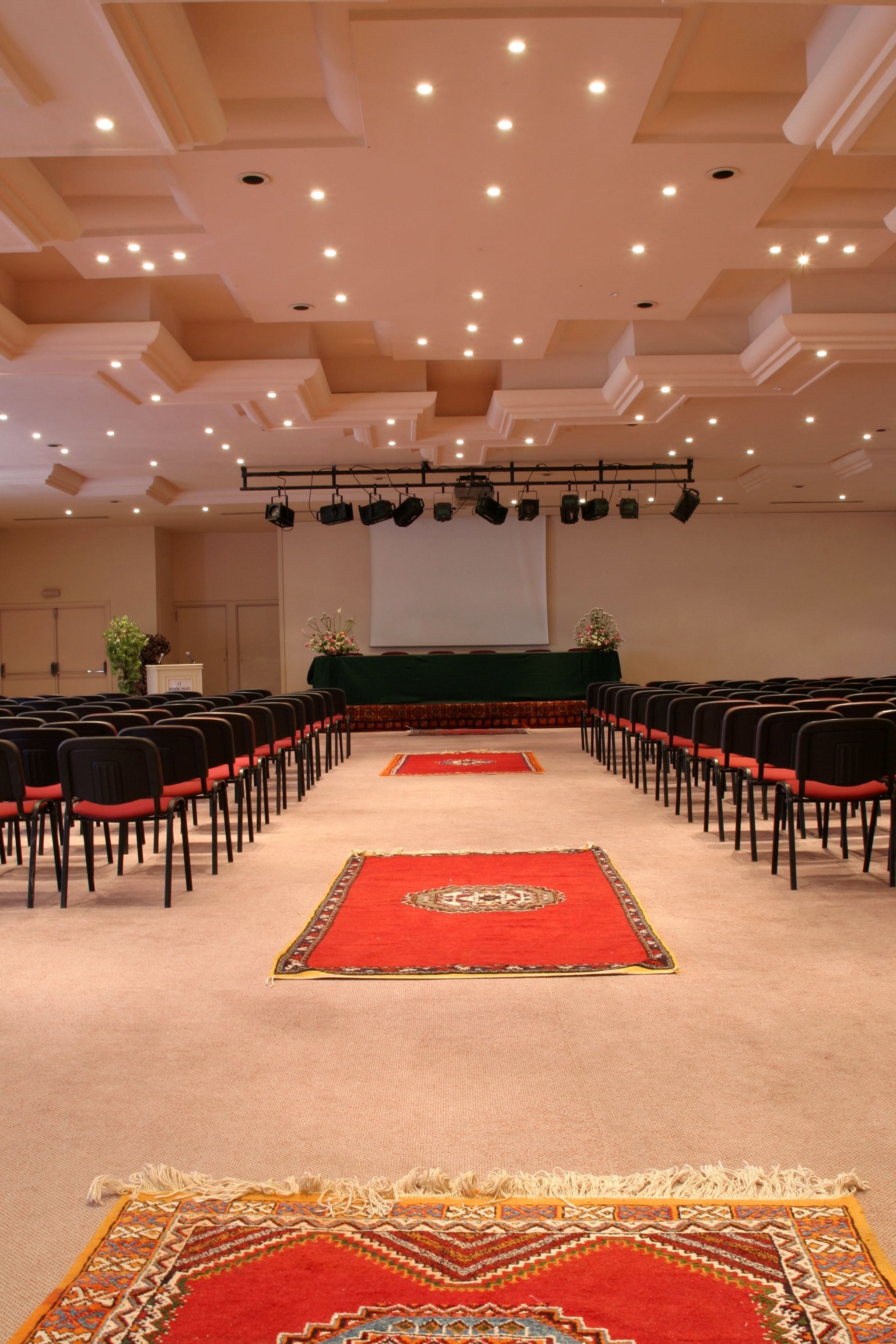 A conference room with rows of chairs, a stage with a projector screen, and a carpeted floor.