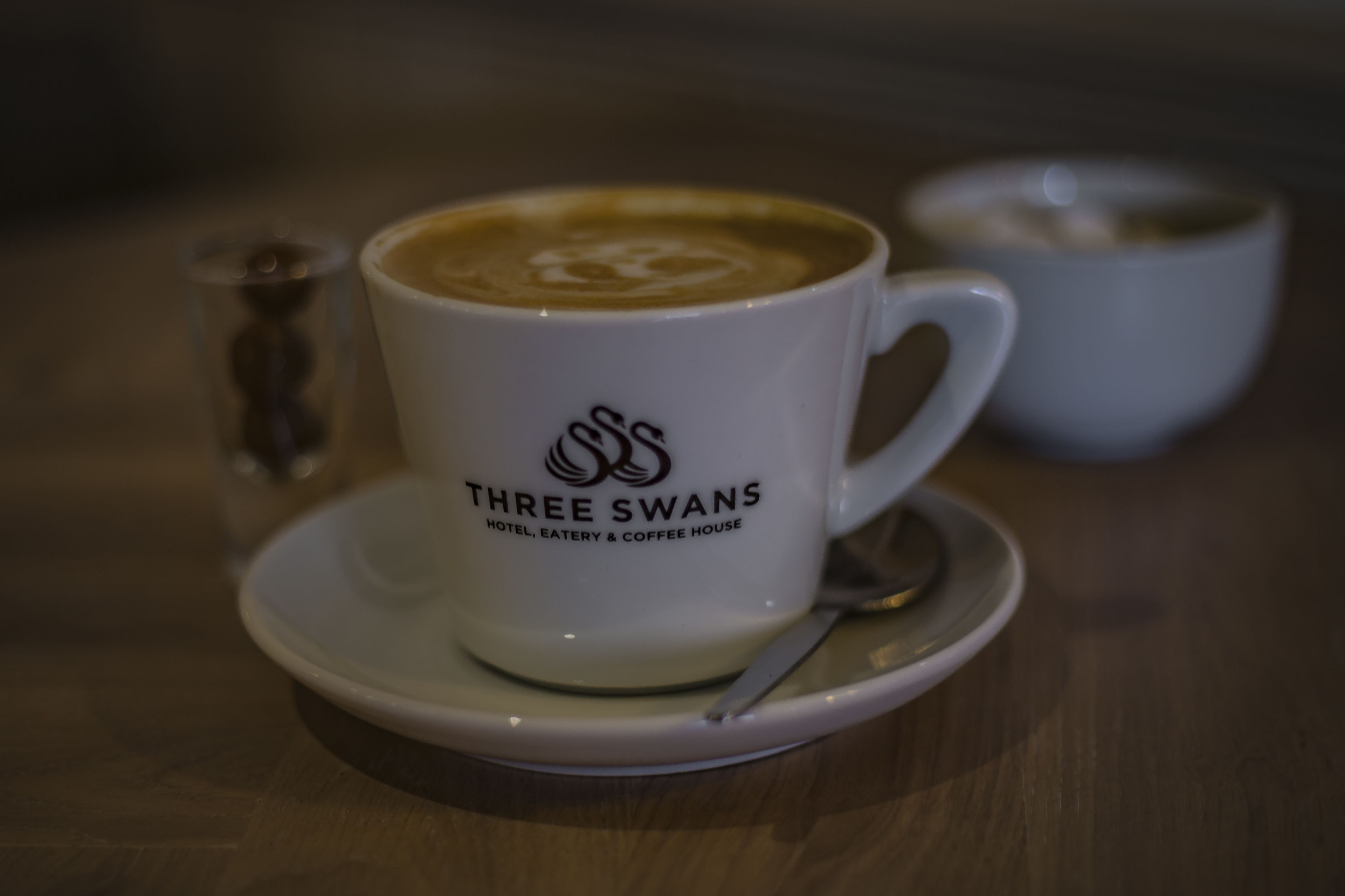 Una taza blanca con el logotipo de 'Three Swans Hotel, Eatery & Coffee House', una cuchara y un plato sobre una mesa de madera.