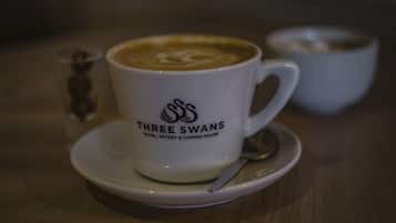 Una taza blanca con el logotipo de 'Three Swans Hotel, Eatery & Coffee House', una cuchara y un plato sobre una mesa de madera.