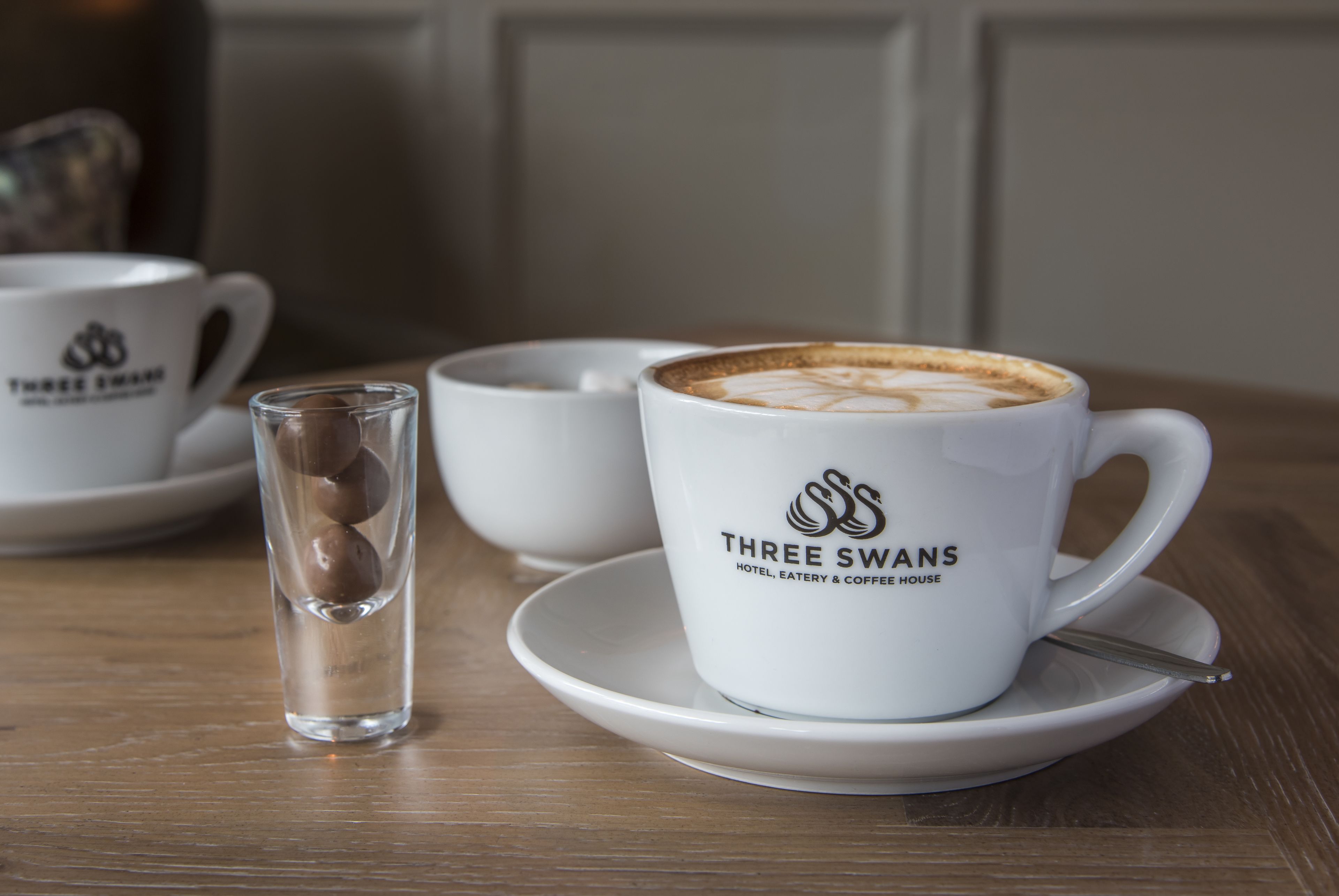 Eine weiße Tasse mit dem Schriftzug "Three Swans" auf einem Untertasse, ein Glas mit einem braunen Gegenstand und eine weitere Tasse im Hintergrund.